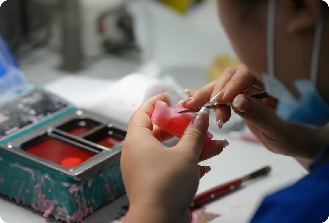 Close-up of hands sculpting or carving a pink wax model with a small tool in a workshop setting.