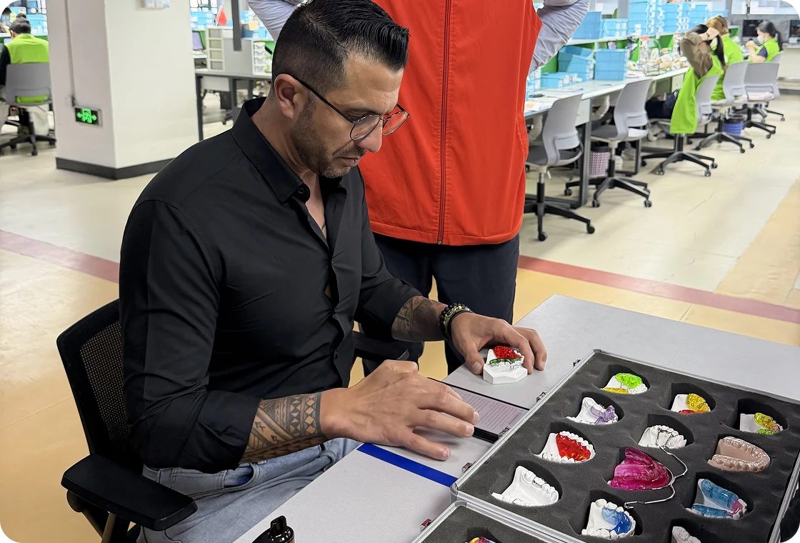 Dr. Sanad Al Murayati in black shirt examining dental molds and orthodontic retainers at a workstation in a factory or lab setting.