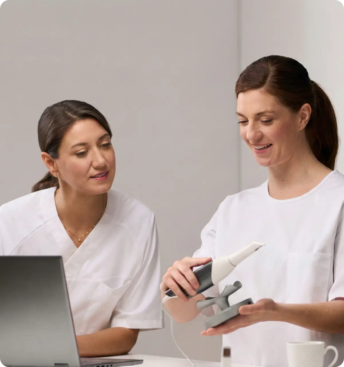 Two female healthcare professionals in white uniforms, one holding and demonstrating a medical device while the other watches attentively near a laptop.