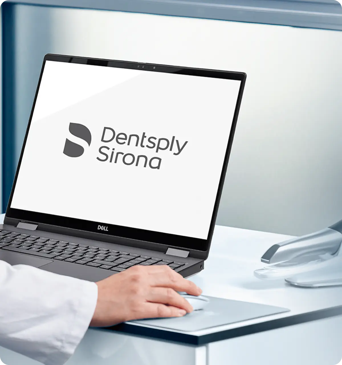 Hand using a mouse next to a Dell laptop displaying the Dentsply Sirona logo on the screen.