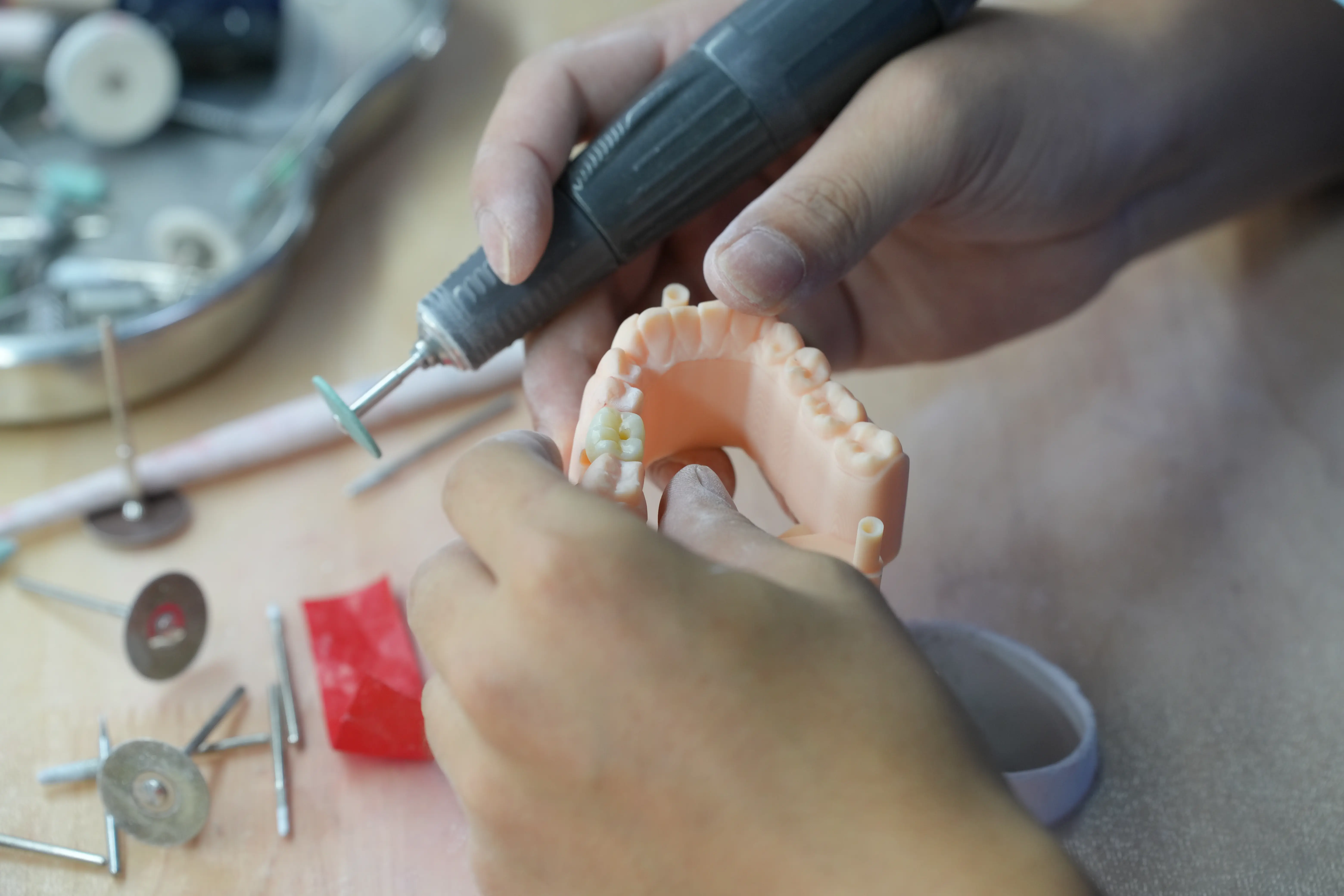 Hands holding a dental drill and working on a dental mold with surrounding dental tools on the table.
