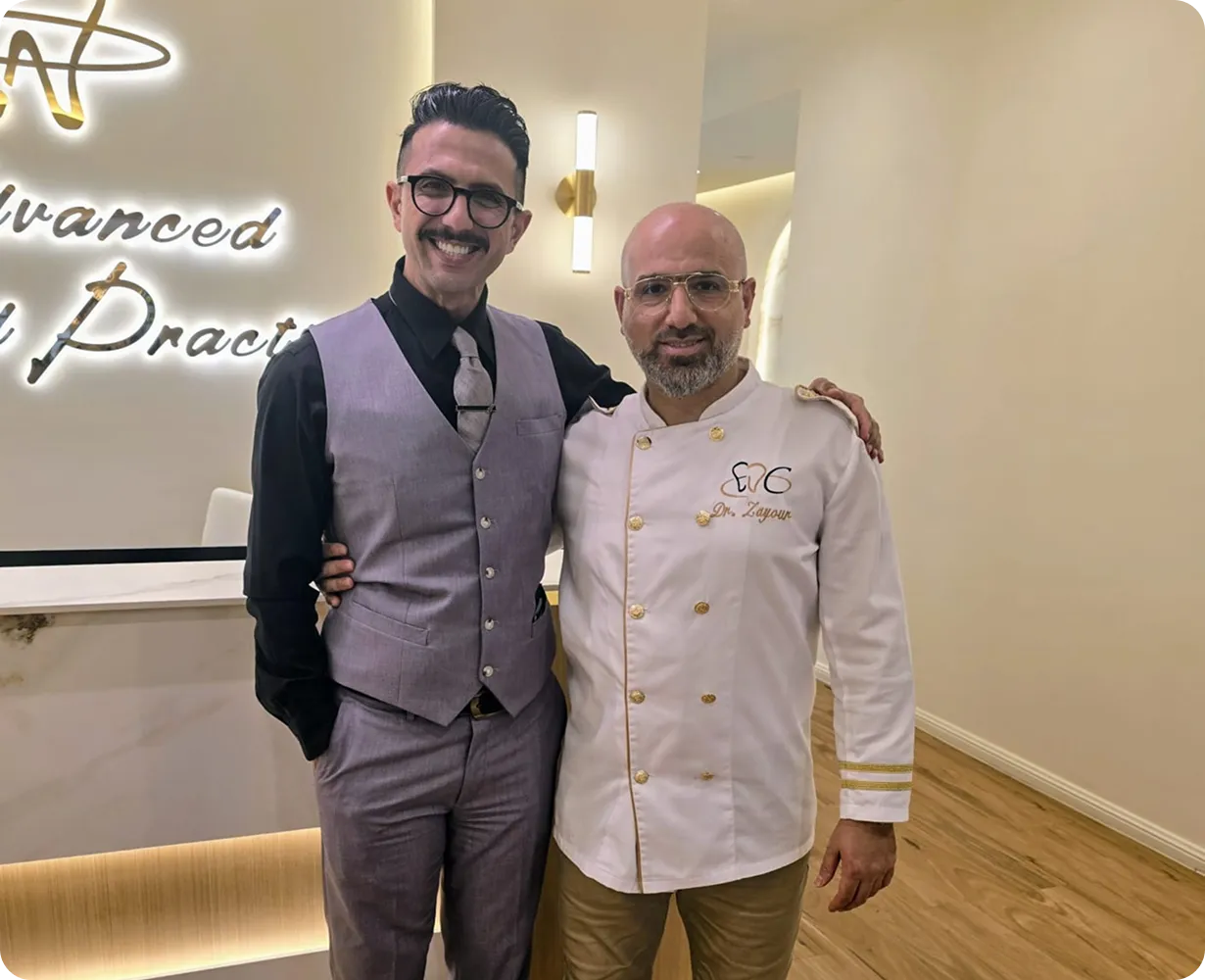 Two men standing indoors; one in a gray vest and tie smiling with glasses, the other in a white medical coat embroidered with 'Dr. Zayour', both posing arm-in-arm.