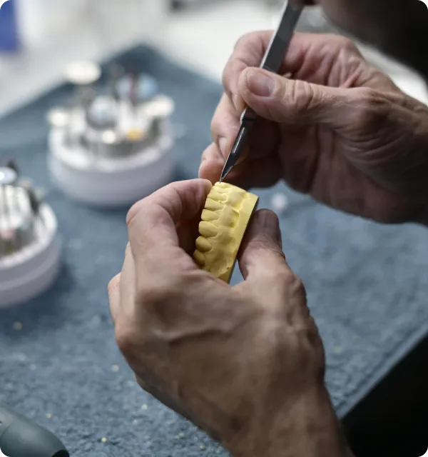 Hands crafting a yellow dental mold using a metal tool on a gray surface.