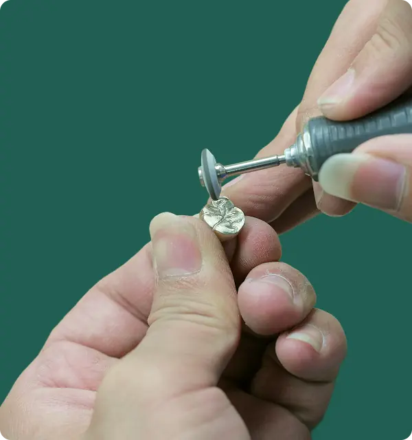 Hands holding and polishing a flower-shaped metal object with a rotary tool against a green background.