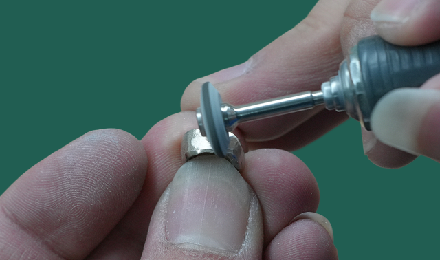 Hands holding and polishing a metal ring with a rotary tool against a green background.