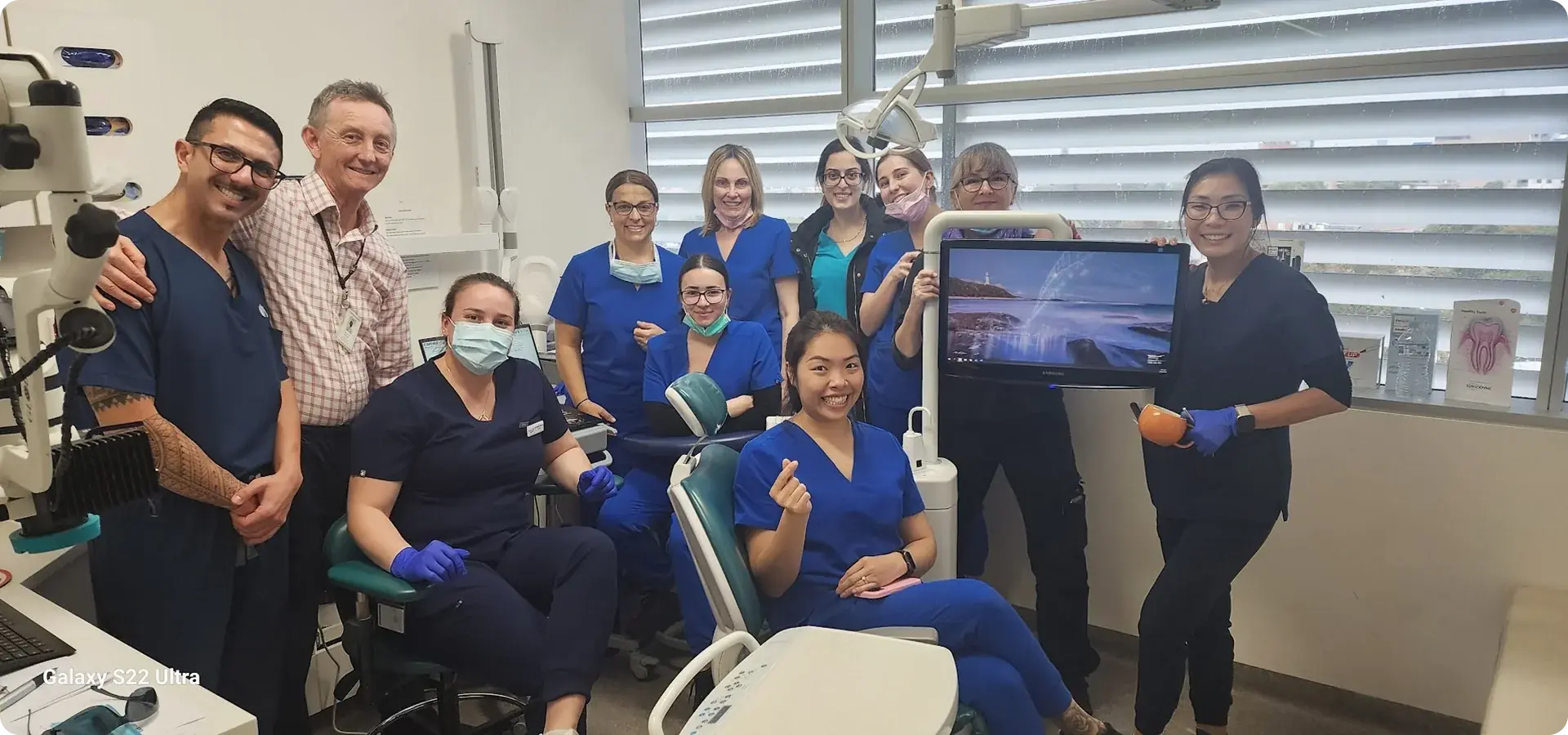 Group of dental professionals in scrubs posing together in a dental office around a patient chair and dental equipment.