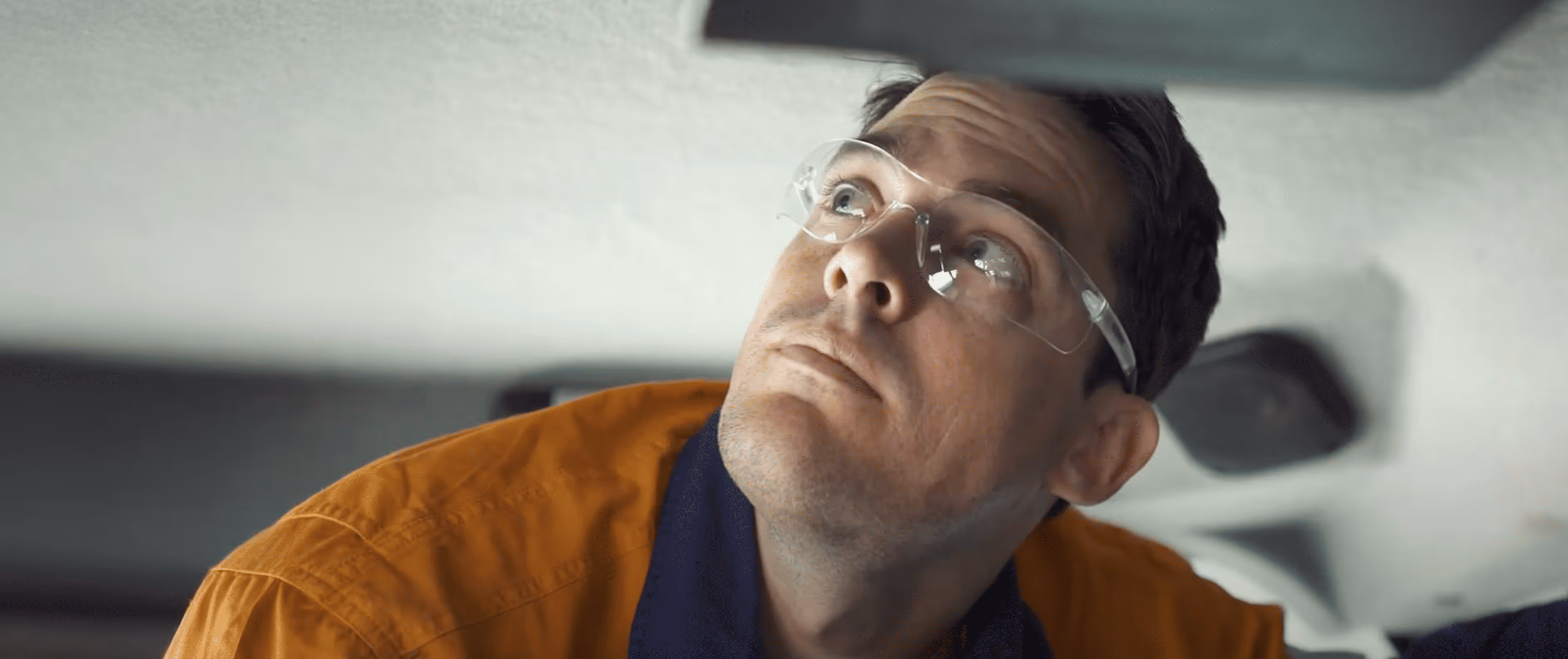 Man wearing safety glasses and an orange shirt looking upward with a focused expression.