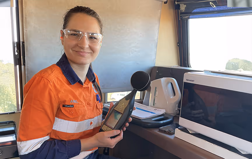 Woman in safety glasses and orange work uniform holding a sound level meter inside a room with a microwave and electric kettle.