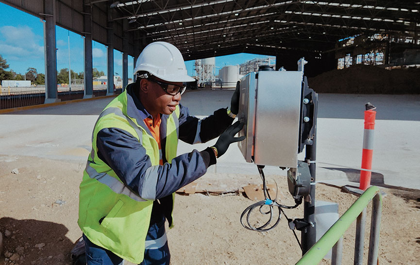 Construction worker in a high-visibility vest and hard hat operating industrial equipment under a large roofed structure.