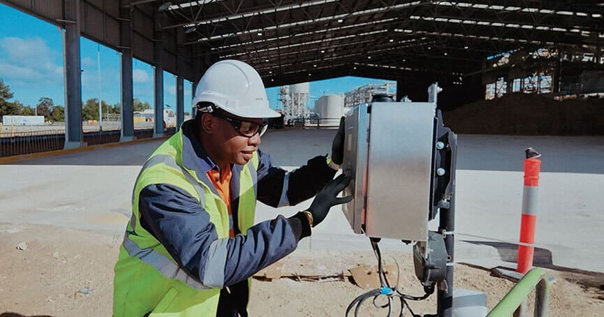 Worker wearing a safety helmet, glasses, gloves, and a high-visibility jacket adjusting a control box in an open industrial warehouse.