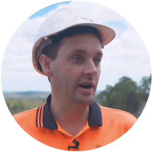 Man wearing a white hard hat and orange Hanson safety shirt speaking outside with trees and sky in the background.