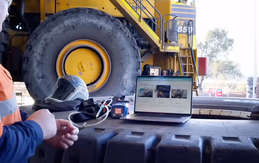 A person in a safety vest near a large industrial vehicle tire, a laptop, and a safety helmet on a workbench.