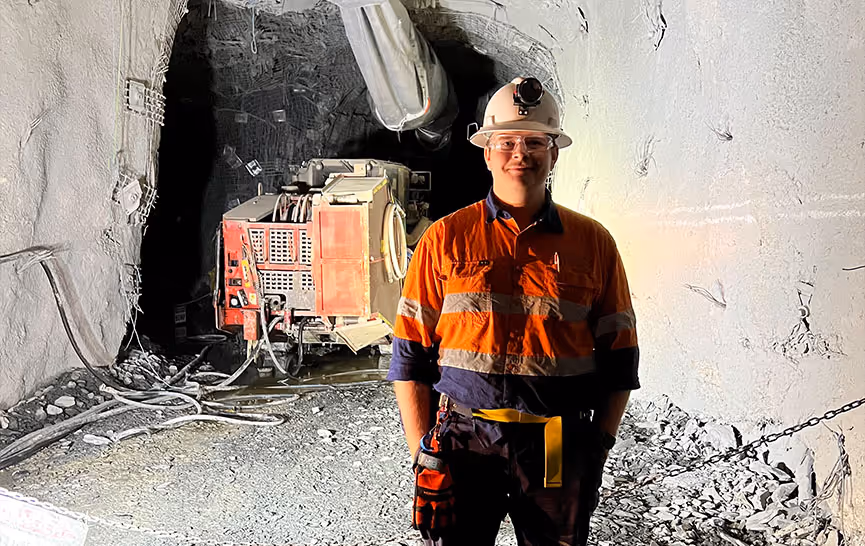 Miner in orange safety gear and helmet standing inside a rocky underground mine tunnel with machinery in the background.