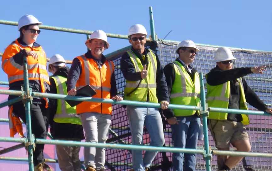 Five construction workers wearing hard hats and high-visibility vests standing on scaffolding under clear blue sky.