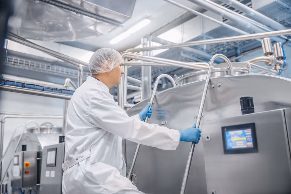 Worker in white protective clothing and hairnet operating stainless steel industrial machinery in a clean factory setting.