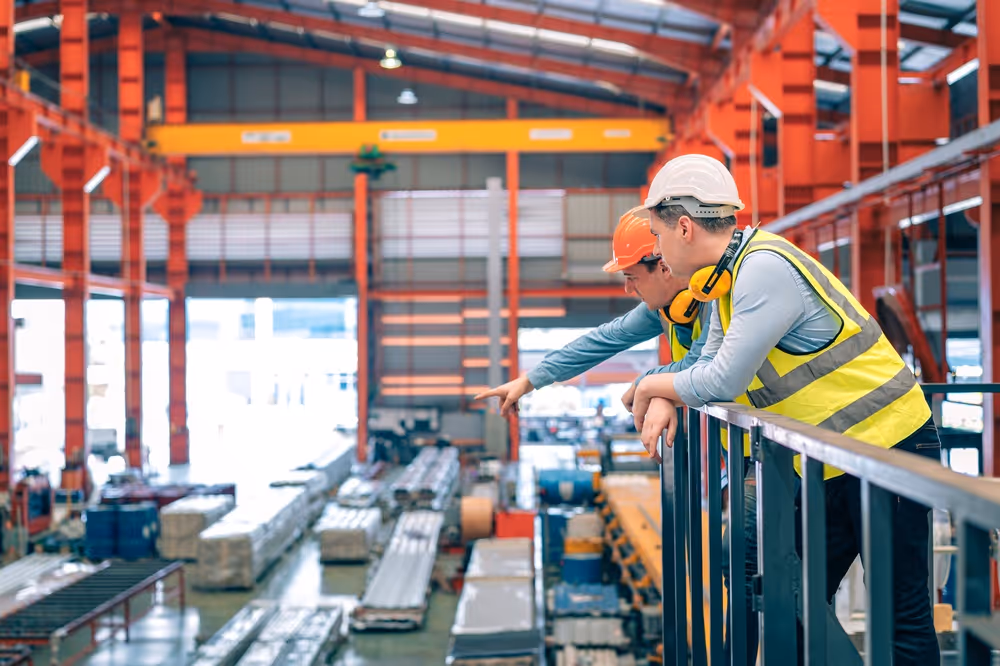 Two construction workers wearing safety helmets and vests look and point at materials inside a large industrial warehouse.