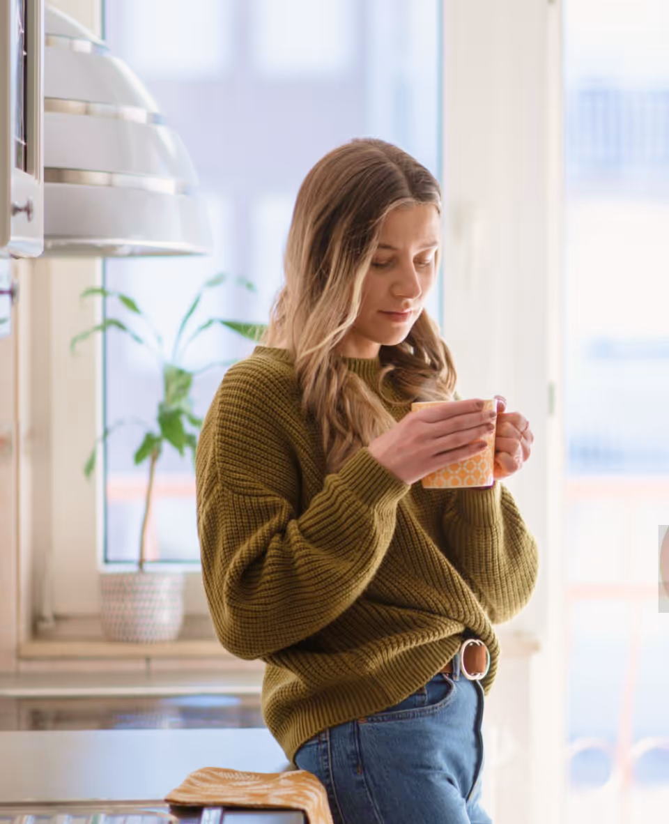 Woman in a green sweater holding a yellow patterned mug in a bright kitchen.