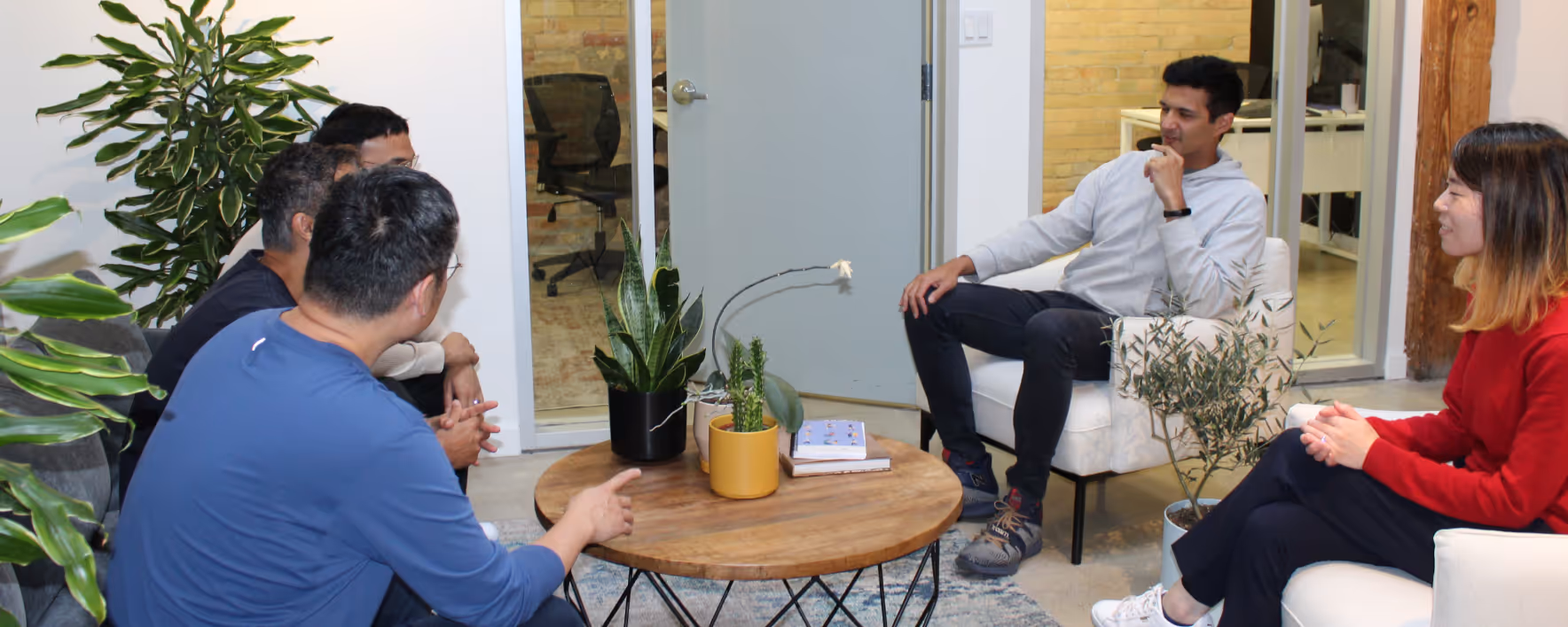 Five people having a casual meeting in a modern office lounge with a round wooden table and indoor plants.