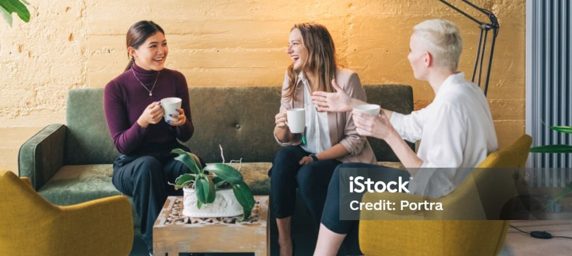 Three women sitting in a cozy room, chatting and smiling while holding white mugs.