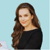 Smiling young woman with long brown hair wearing a black top against a light blue background.