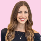 Smiling woman with long wavy brown hair wearing a black top and a necklace against a pink background.