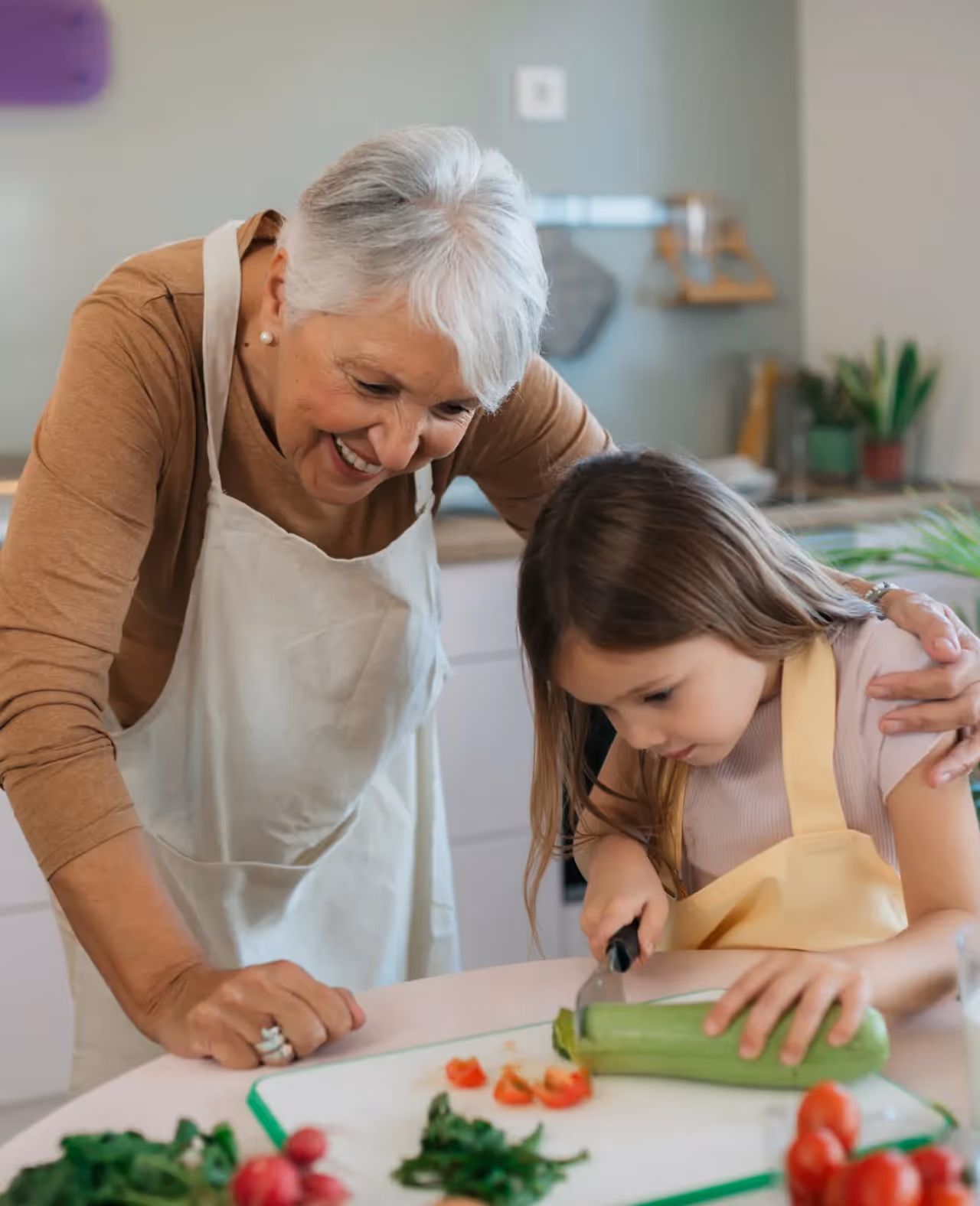 Smiling elderly woman watching young girl cut a zucchini in a kitchen.