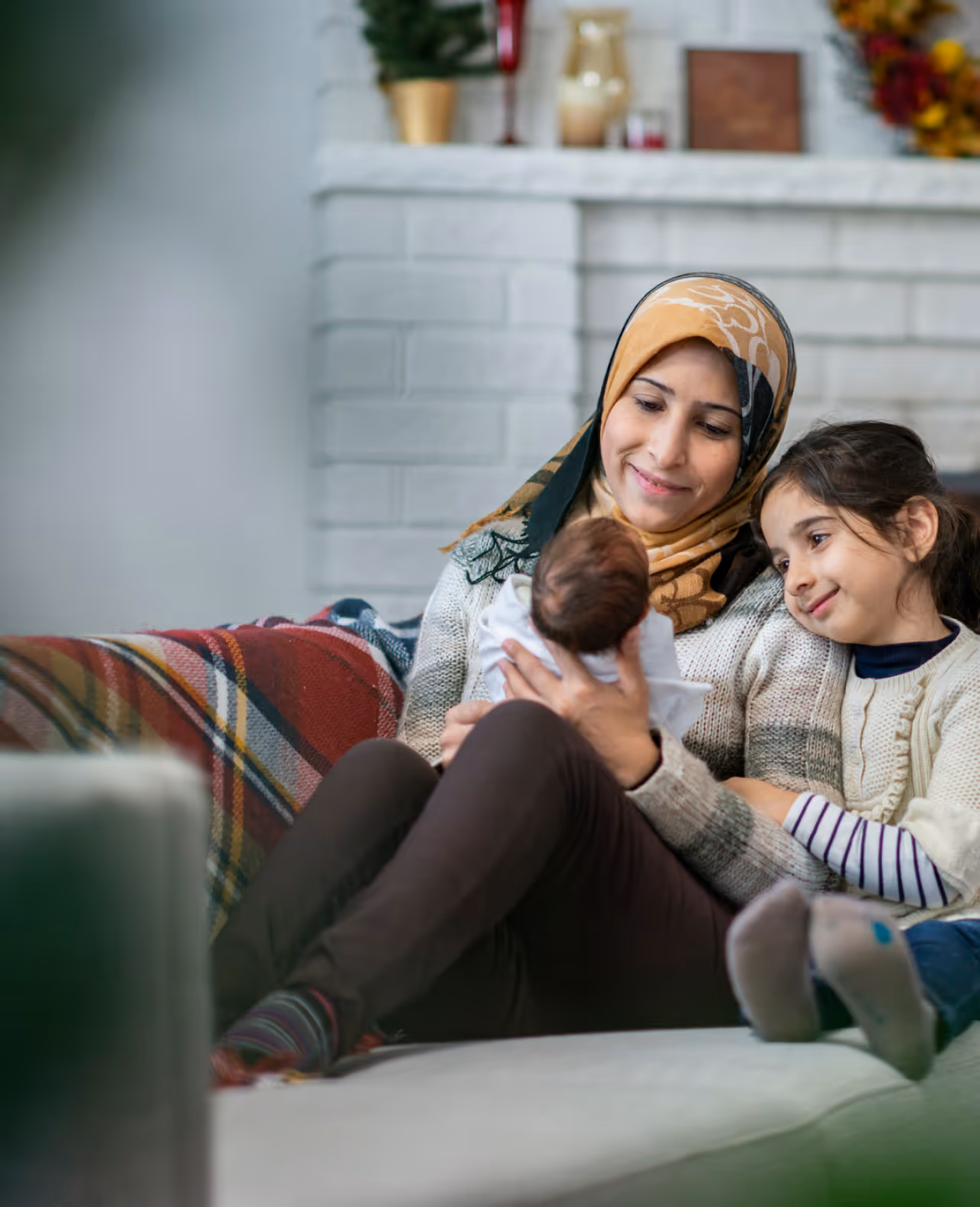 Smiling mother wearing a hijab sitting on a couch holding a newborn baby, with a young girl leaning on her shoulder.