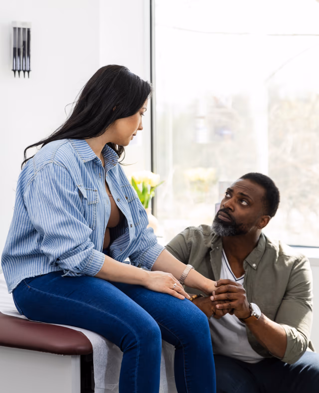 Man holding and comforting a seated woman in a room with a large window.