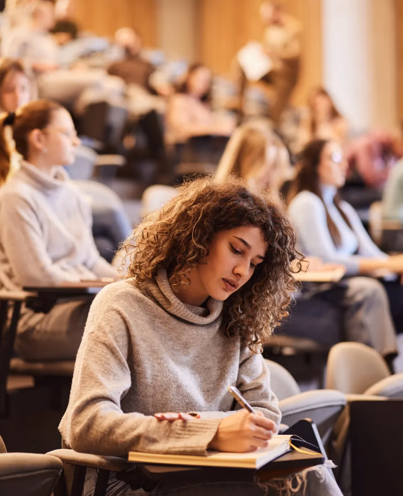 Young woman with curly hair writing in a notebook in a lecture hall with other students seated behind her.