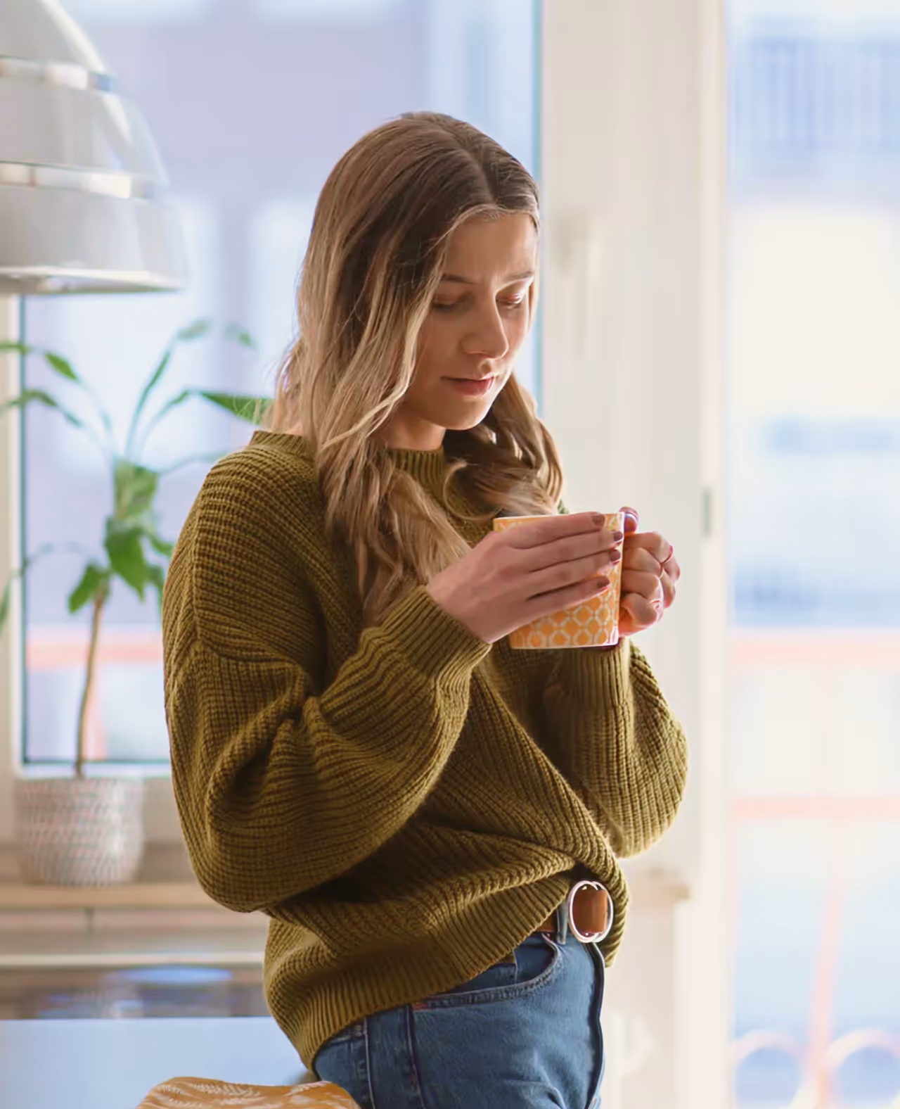 Woman in olive green sweater holding and looking at a yellow patterned mug in a bright room.
