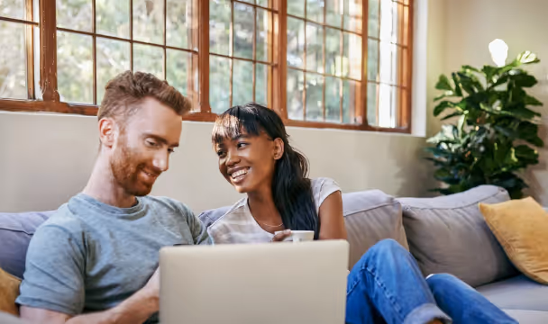Smiling couple sitting on a couch looking at a laptop together in a sunlit room.