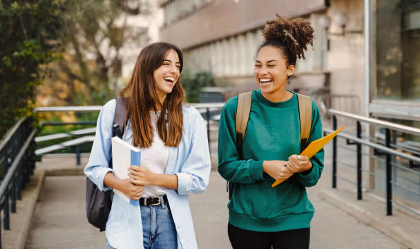 Two young women with backpacks smiling and talking while walking outside on a campus pathway.