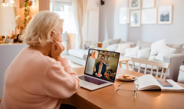 Elderly woman sitting at a table having a video call on a laptop with a man, with books and cookies on the table.