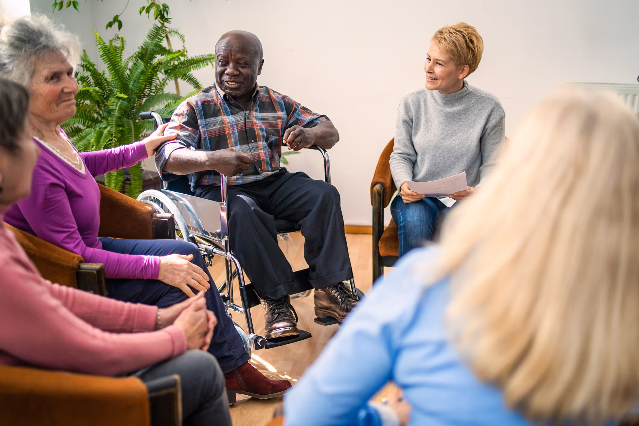 A diverse group of older adults sitting in a circle with a therapist in a bright room, engaging in a supportive conversation.