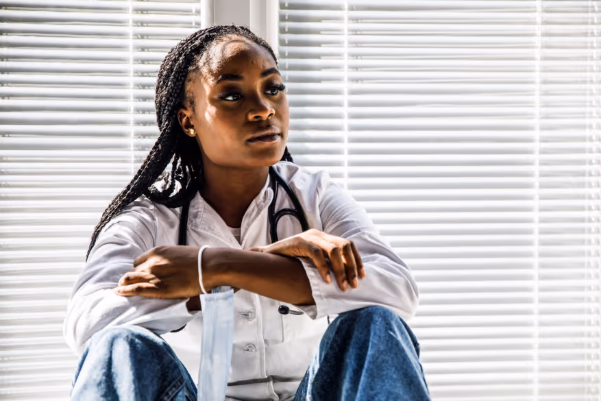 Thoughtful female doctor in white coat with stethoscope sitting with arms crossed, holding a face mask.