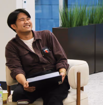 Smiling man sitting on a chair holding a laptop in a modern indoor setting with plants in the background.