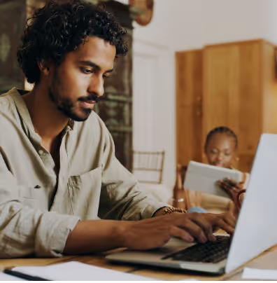 Man with curly hair typing on a laptop while a woman reads a book in the background.