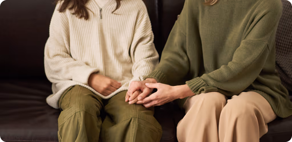 Two people sitting on a dark couch, one wearing a cream sweater and olive pants, the other in a green sweater and beige pants holding the other's hand.