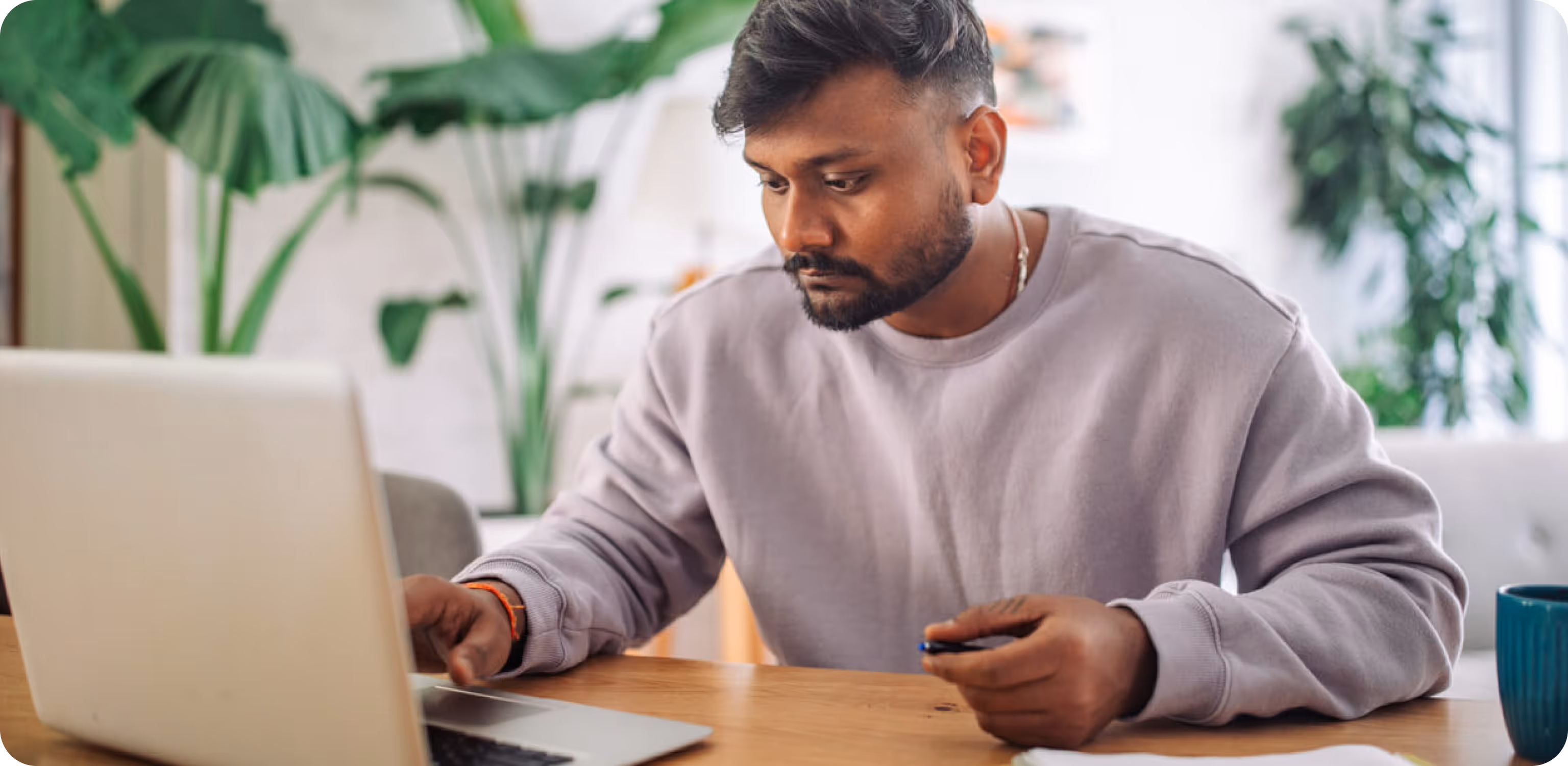Man in a light purple sweatshirt working intently on a laptop at a wooden table in a room with green plants.