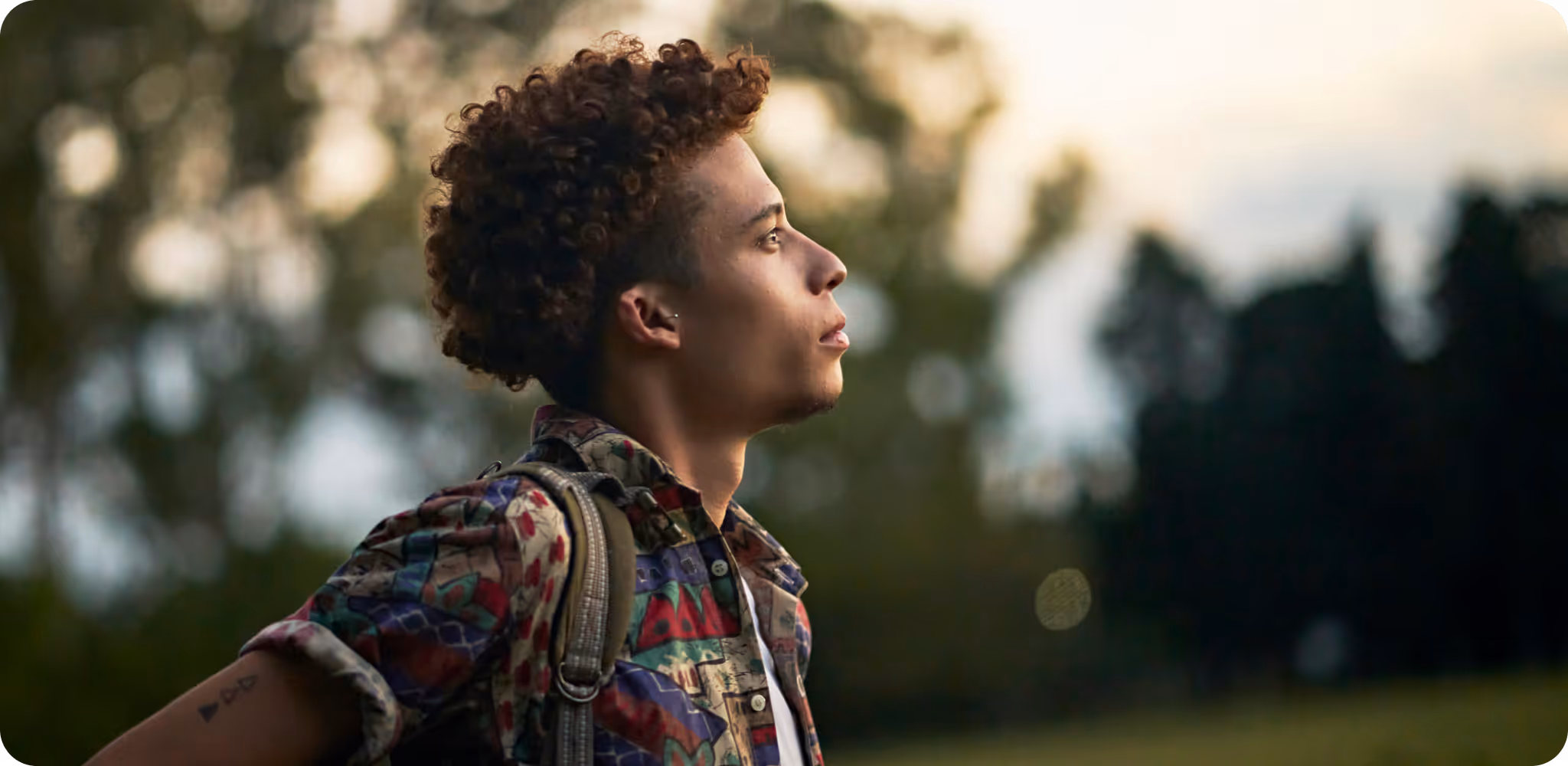 Young man with curly hair and a patterned shirt looking thoughtfully into the distance outdoors at sunset.