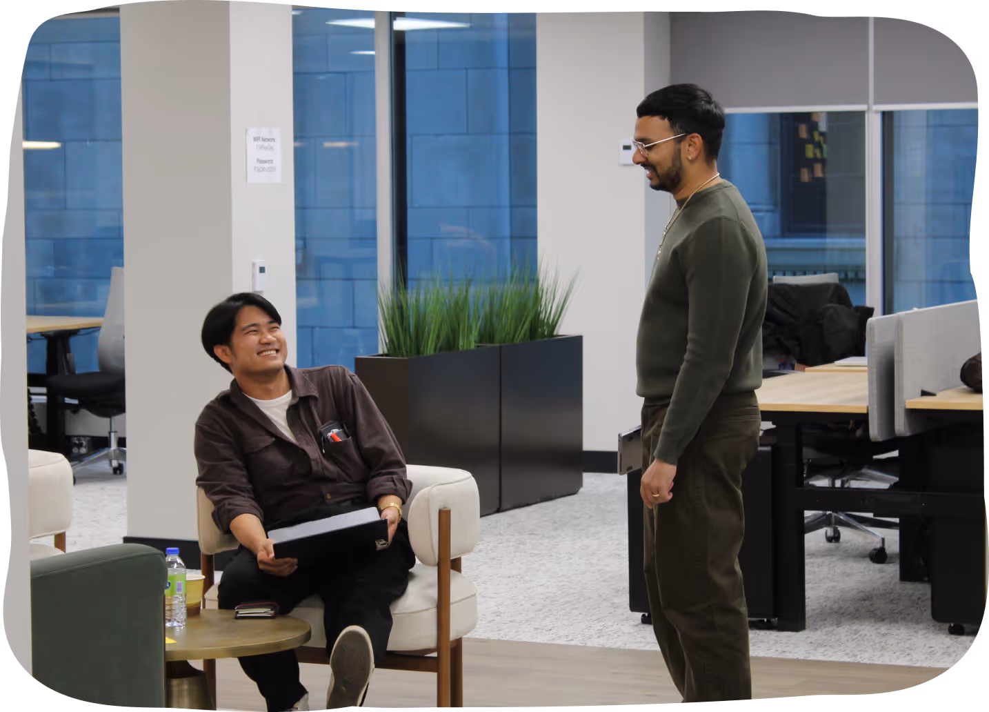 Two men smiling and interacting in a modern office lounge area with chairs and plants.