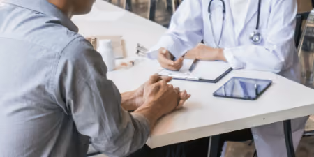 Patient consulting with a doctor at a desk with a tablet and clipboard.