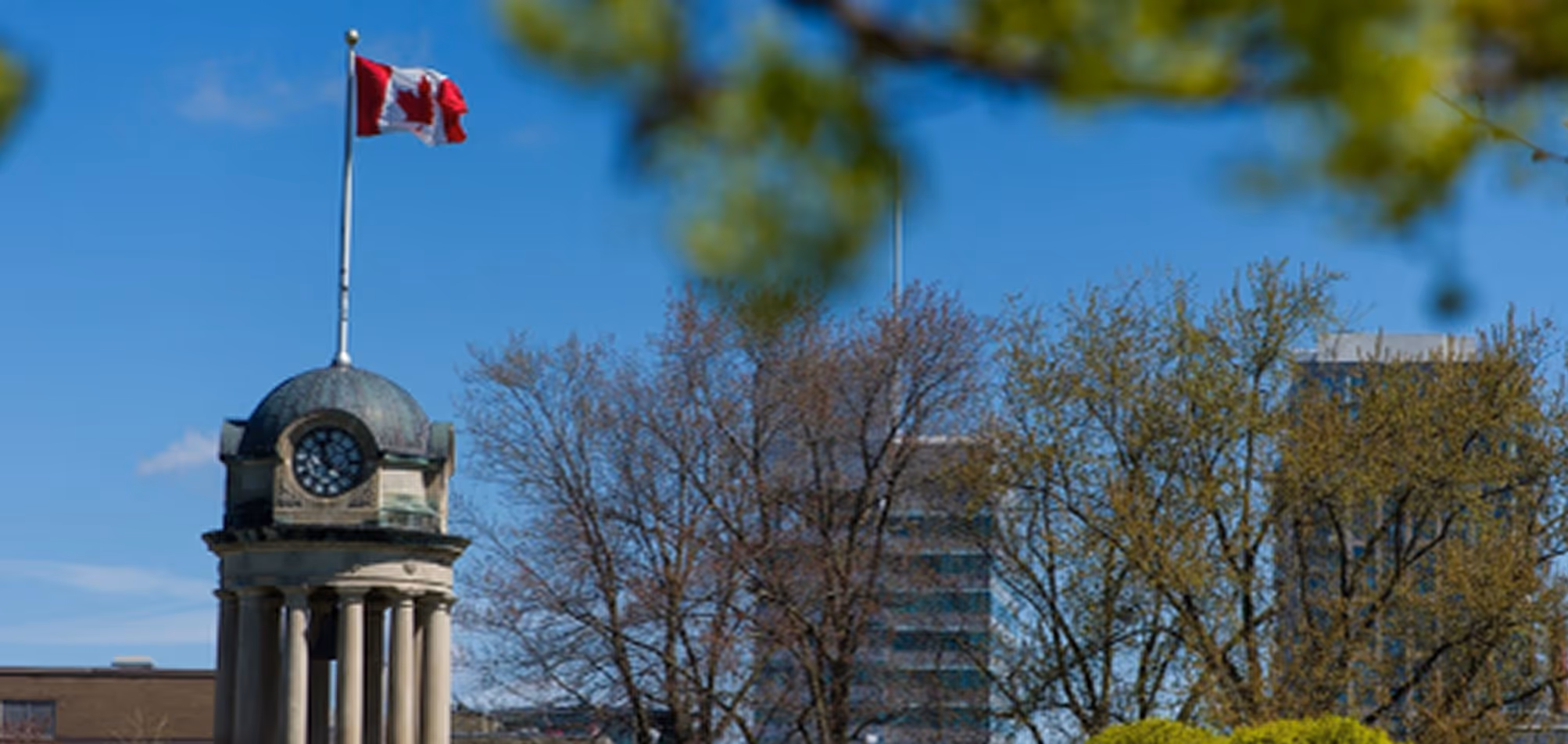 Canadian flag flying atop a clock tower with trees and buildings in the background under a clear blue sky.