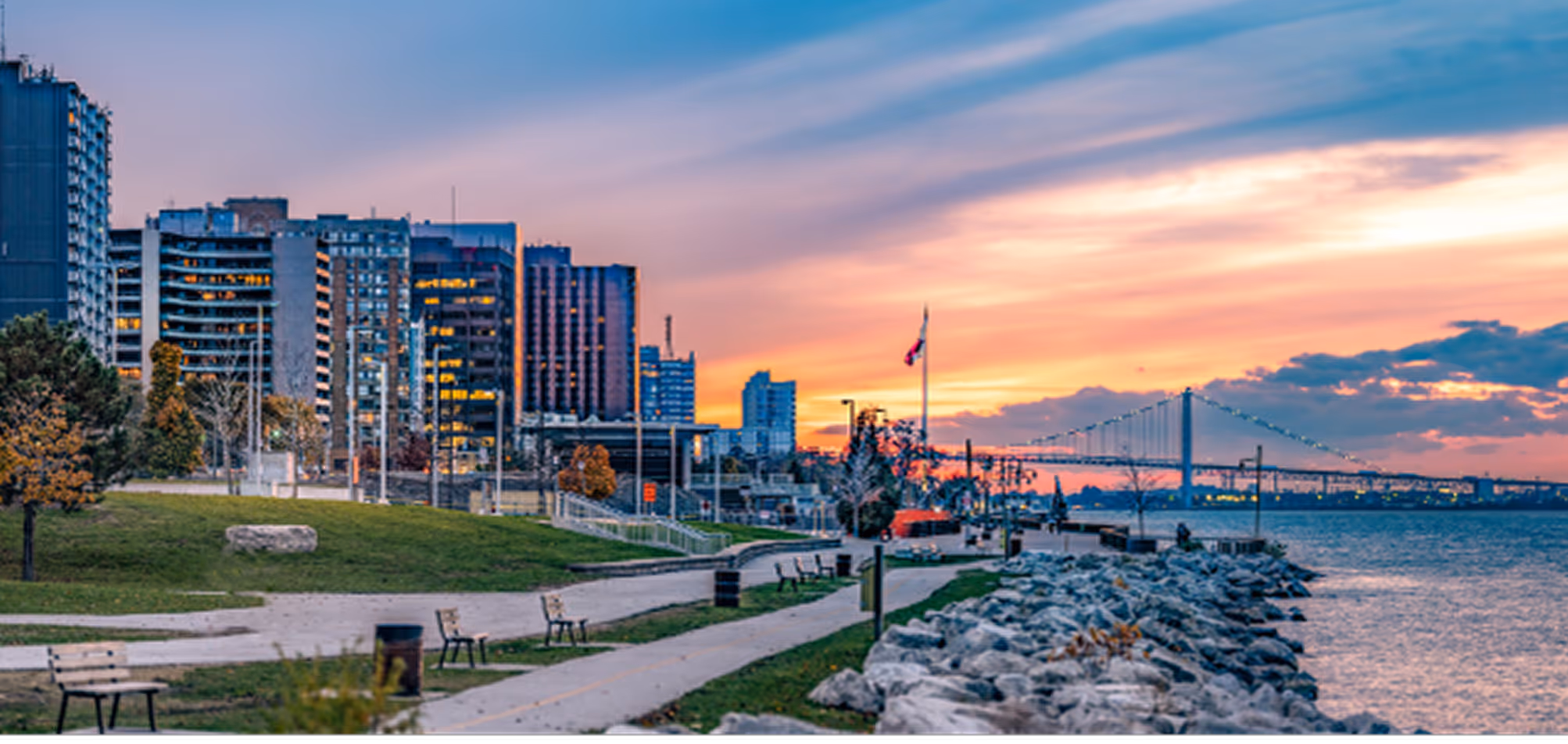Sunset over waterfront park with benches, city buildings, and a suspension bridge in the distance.