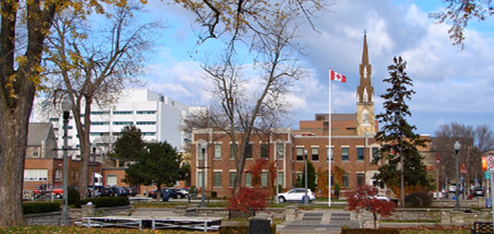 Urban park with trees and Canadian flagpole in front of brick buildings and church steeple.