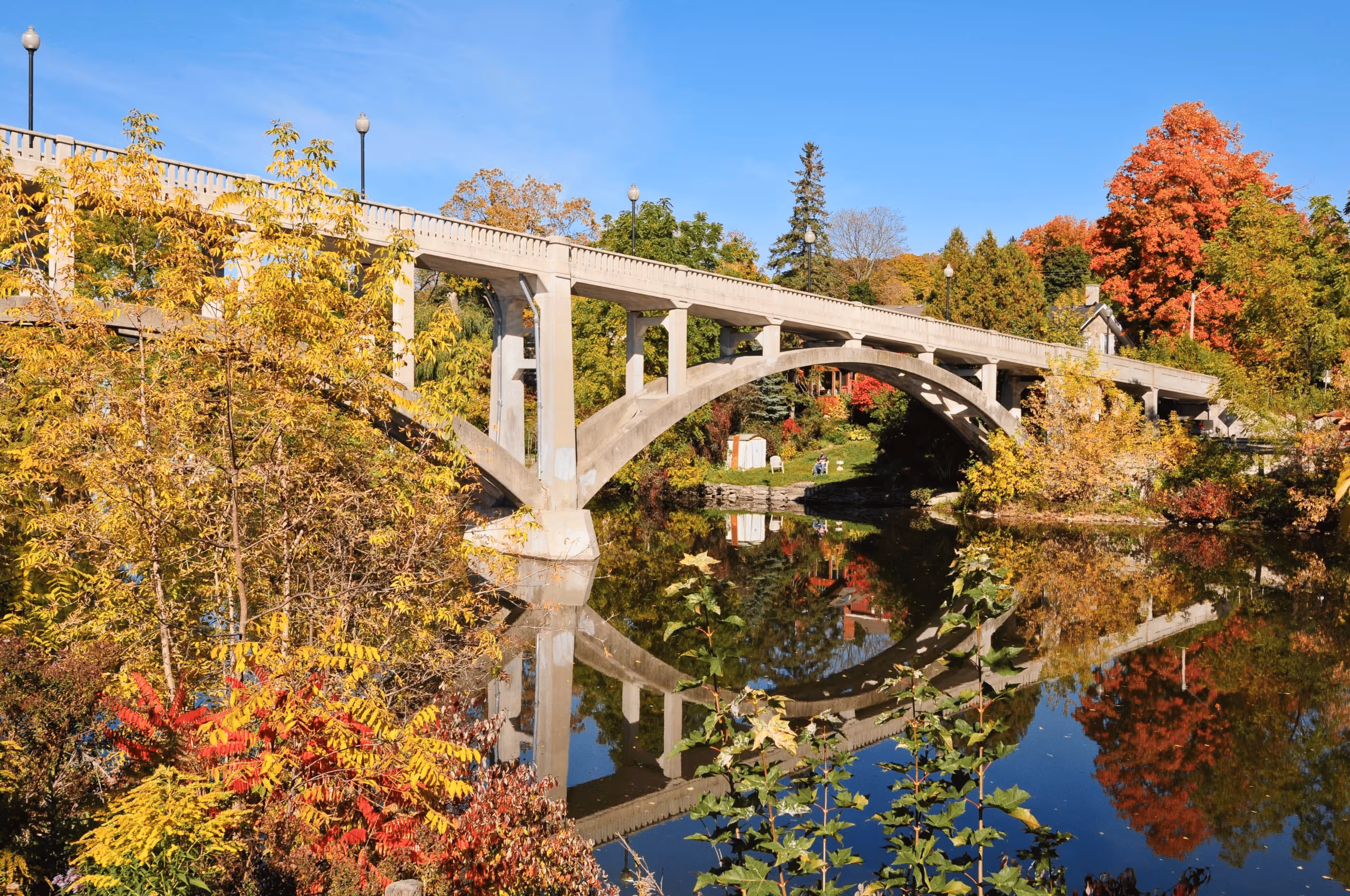 Concrete arch bridge over calm river reflecting colorful autumn trees under a clear blue sky.