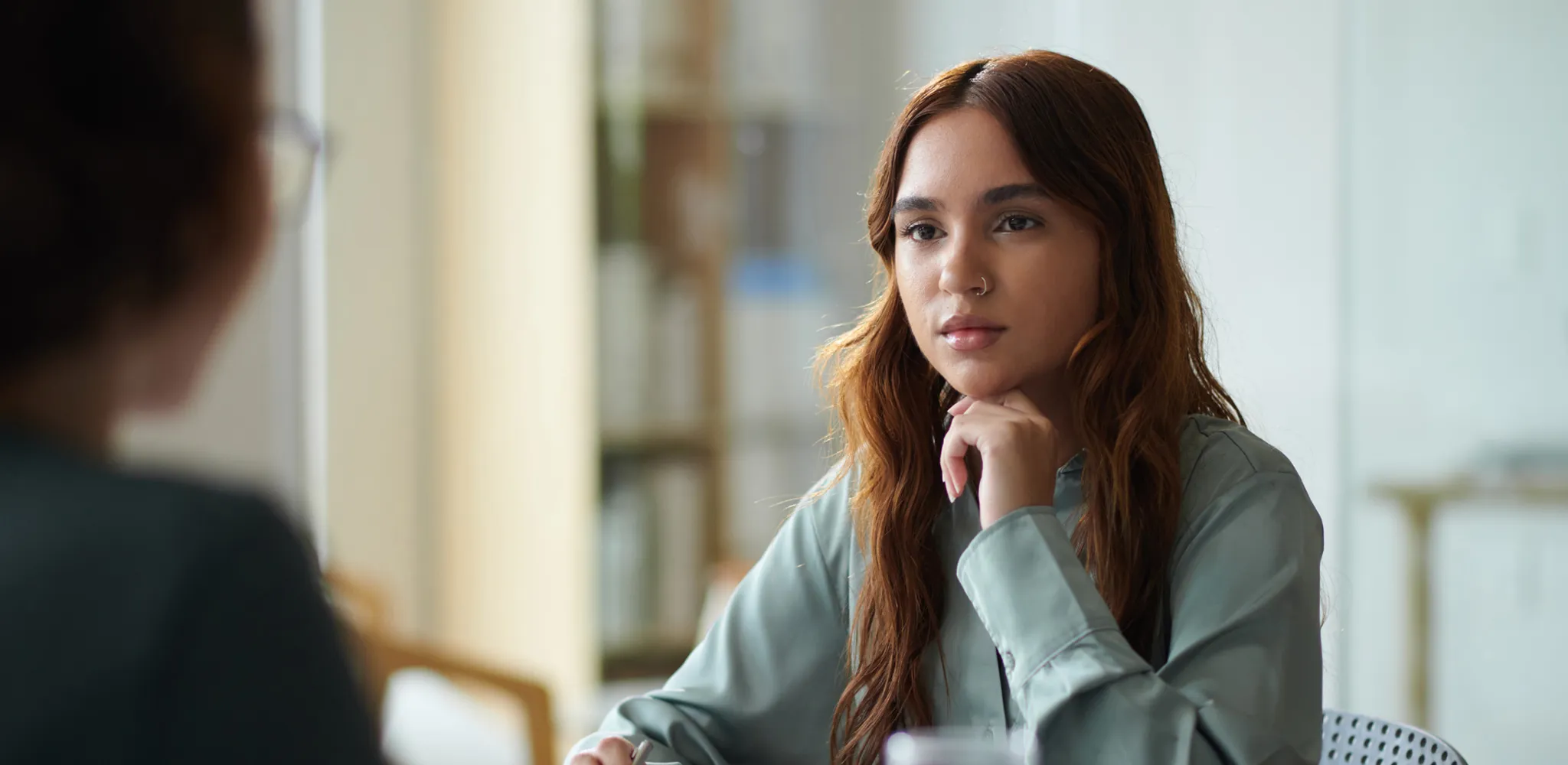 Young woman with long brown hair and a nose ring, wearing a light blue shirt, sitting at a table with her chin resting on her hand, attentively listening to another person whose blurred silhouette is visible in the foreground.