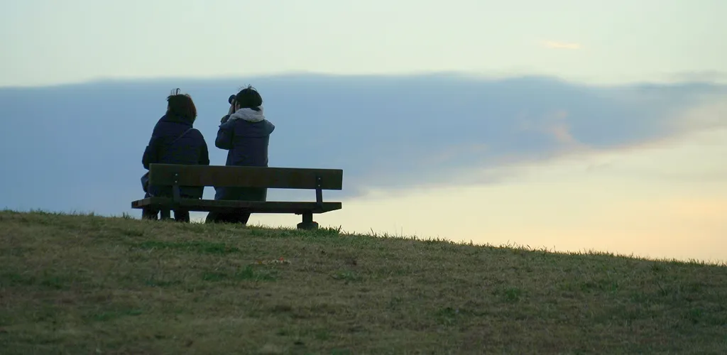 Two people sitting on a wooden bench on a grassy hill during sunset, one looking through binoculars toward the horizon.