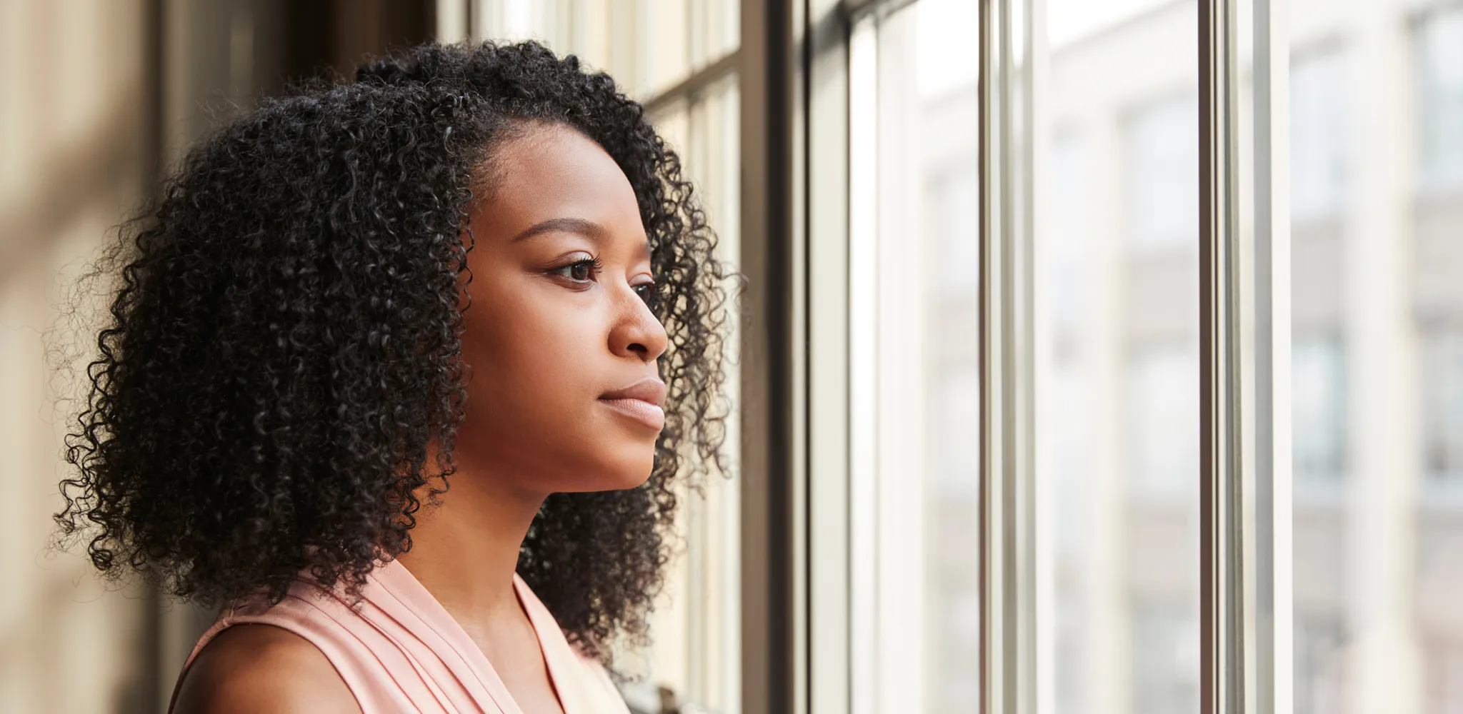 Side profile of a young woman with curly hair looking thoughtfully out a large window with buildings visible outside.