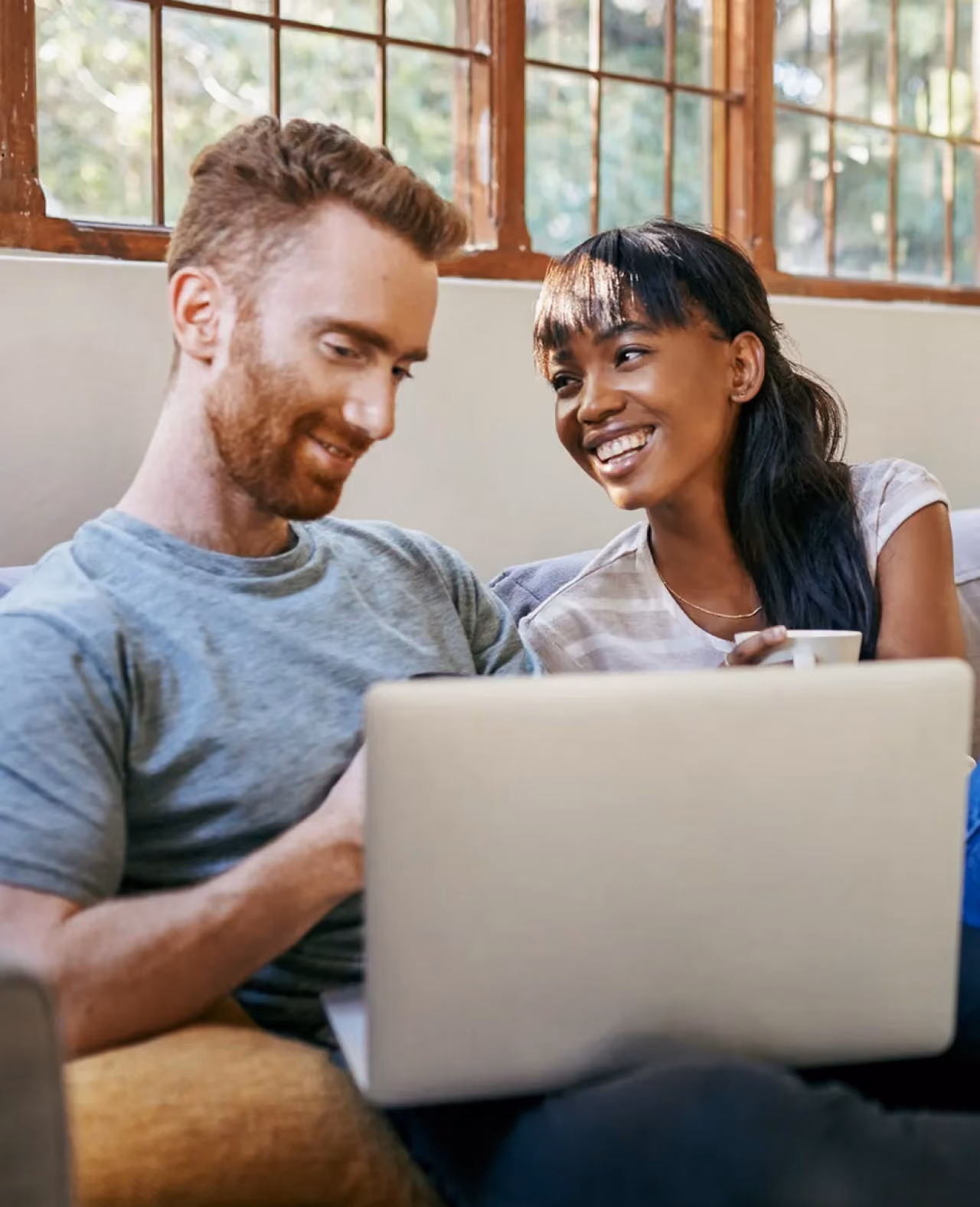A smiling couple sitting closely together on a couch, looking at a laptop screen with natural light coming through large windows behind them.